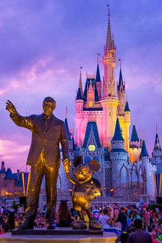 a statue of walt and mickey mouse in front of a castle at dusk with people standing around