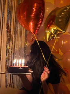 a woman holding a cake with lit candles in front of balloons and streamers on the wall