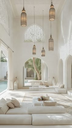 a living room filled with lots of white furniture and hanging lights above the couches