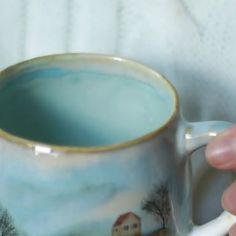 a hand holding a coffee mug with a house on it and trees in the background