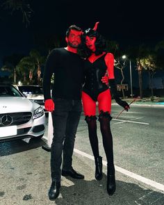 a man and woman dressed up in devil costumes standing next to a white car at night