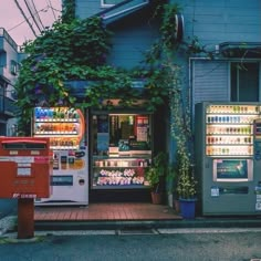 two vending machines sitting on the side of a road next to a building with vines growing over it