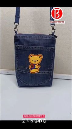 a small denim bag with a teddy bear on the front and side pocket, hanging from a metal hook