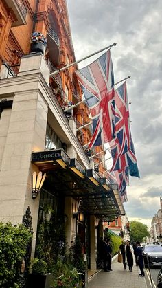 people are walking down the sidewalk in front of a building with flags flying from it