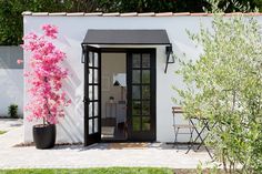 a small white house with black doors and pink flowers in the potted planter