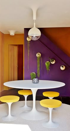 a white table and four yellow chairs in front of a purple wall with plants on it