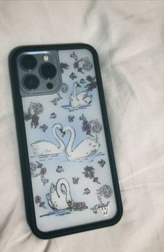 an iphone case with swans on it sitting on a white bed sheet, next to two black buttons