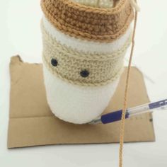 a knitted coffee cup sitting on top of a piece of cardboard with a needle in it