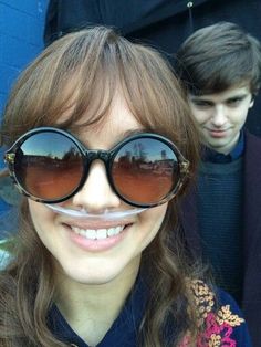 a woman wearing large round sunglasses with fake moustaches on her face, and another man in the background