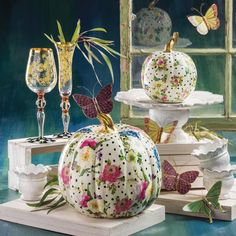 three white pumpkins decorated with flowers and butterflies sit on pedestals next to wine glasses