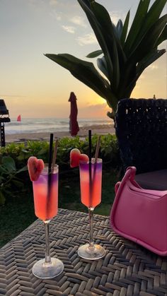 two glasses with drinks sitting on top of a table next to the ocean at sunset