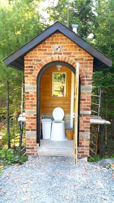 a toilet in a small brick building surrounded by trees and graveled area with steps leading up to it