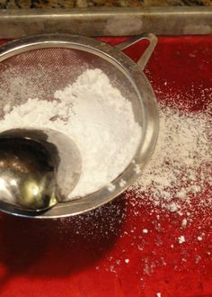 a metal strainer filled with white powder on top of a red counter next to a silver spoon