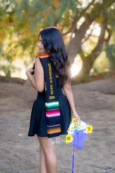 a woman in a black dress holding a bouquet of flowers and a colorful sash around her neck