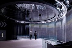 two men are standing in front of a circular glass wall with lights hanging from the ceiling