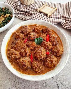 meatballs and spinach are in a red sauce on a white plate next to a bowl of greens