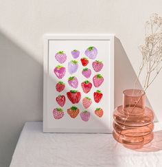 a pink vase with strawberries on it next to a white wall and a framed photograph