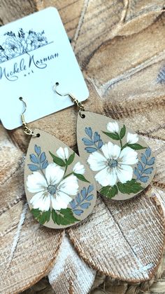 the earrings are decorated with white flowers and green leaves on wood planks next to a card