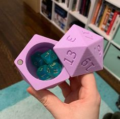 a hand holding a pink and blue dice with numbers on it in front of a bookshelf