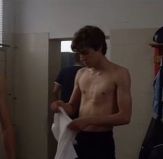 two young men standing in a bathroom looking at their shirts hanging on a rack and one man is not wearing a shirt