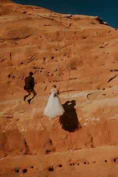 a bride and groom are climbing up the side of a mountain with their shadow on the ground