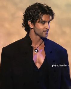 a man with curly hair wearing a black suit and necklace in front of a brown background