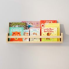 A natural wood wall shelf filled with children's books, mounted on a light gray wall. The shelf holds colorful book covers, making it an inviting and organized space for young readers. Soft Toy Storage, Floating Bookshelf, Nursery Bookshelf, Kids Shelves, Nursery Trends, Wall Mounted Bookshelves, Nursery Shelves, Wood Bookshelves, Prop Stylist