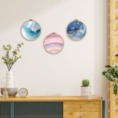 three circular paintings hang on the wall above a dresser in a white room with blue and pink accents