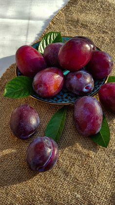 some plums are in a bowl on a table with green leafy leaves and brown burlock