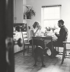 two people sitting at a kitchen table with a dog on the floor in front of them