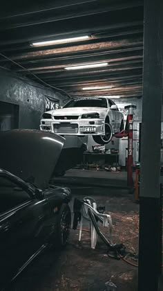 two cars are parked in a garage with their hoods open and one car is being worked on