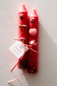 two red candles with flowers and hearts on them are tied to a white wall by a pink ribbon