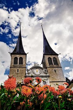 an old church with two steeples and flowers in the foreground against a cloudy blue sky