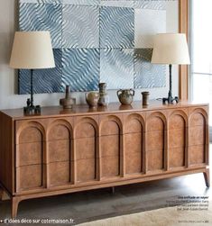 a wooden dresser with two lamps on top of it and an art piece behind it