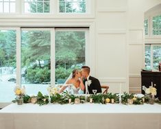 a bride and groom kissing in front of a long table with greenery on it