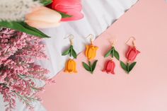 three pairs of flower earrings sitting on top of a pink surface next to tulips