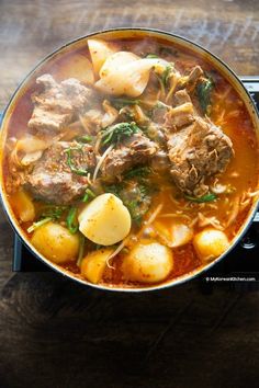 a pot filled with meat and potatoes on top of a stove burner next to a wooden table