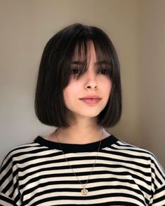 a woman with short black hair wearing a striped shirt and gold necklace, standing in front of a white wall