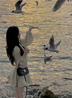 a woman standing on the beach with seagulls flying over her and looking at the water