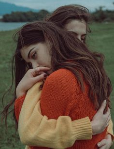 two young women embracing each other in a field with mountains in the background, one woman is wearing an orange and yellow sweater