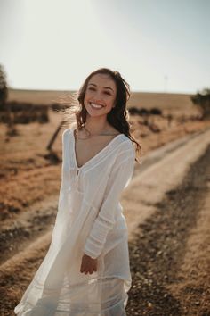 a woman standing in the middle of a dirt road wearing a white dress and smiling