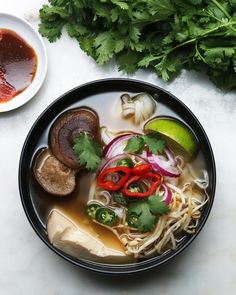 a black bowl filled with soup and vegetables next to some cilantro, lime wedges and sauce