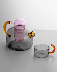 a glass tea pot with an orange and pink handle sitting next to another glass cup