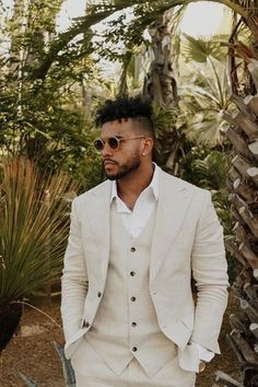 a man wearing a white suit and sunglasses standing in front of palm trees with his hands in his pockets