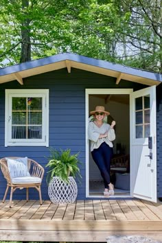 a woman sitting on a porch next to a small blue house with two chairs and a potted plant