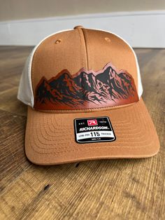 Celebrate the rugged beauty of the mountains with this exquisite Richardson 115 hat featuring the captivating mountain range leather patch. Whether you're exploring nature trails, summiting peaks, or simply enjoying the view, this hat is the perfect companion for your outdoor escapades. This Richardson 115 Caramel/Birch hat featuring a striking leather patch depicting a mountain range has been meticulously matched by our color professionals. This hat and patch combination is perfect for hikers, Rugged Brown Hat For Travel, Rugged Brown Travel Hat, Western Style Snapback Hat For Outdoor, Brown Six-panel Trucker Hat For Outdoor Activities, Adjustable Leather Baseball Cap For Outdoor, Western Style Baseball Cap With Flat Bill For Outdoor, Rugged Leather Hat Bands For Outdoor, Western Hats With Leather Patch For Outdoor, Western Snapback Hat With Flat Brim For Outdoors