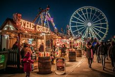 people walking around an amusement park at night with ferris wheel in the background and lights on