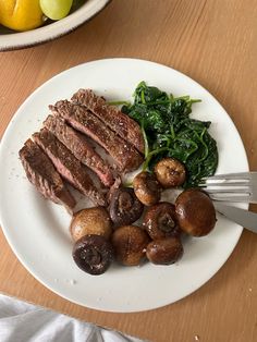 a white plate topped with meat and mushrooms next to a bowl of vegetables on a wooden table