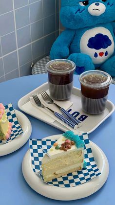 a blue teddy bear sitting at a table with two cups of coffee and a piece of cake