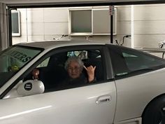 an older woman sitting in the driver's seat of a silver car with her hand up